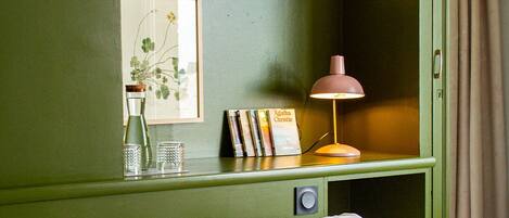 Una habitación con una cama, un escritorio con lámpara, cuadros enmarcados y una estantería llena de libros.