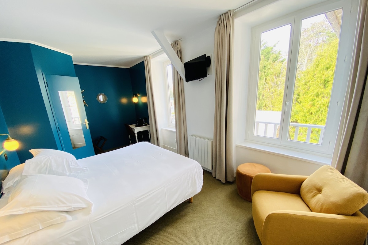 Habitación de hotel con cama, sofá, televisor, ventana y vistas a árboles, y una puerta.