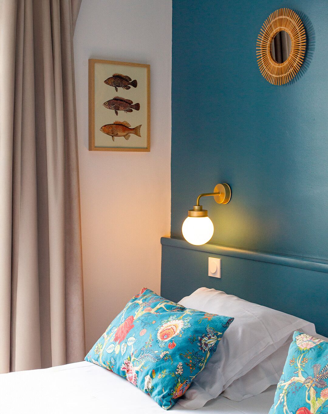 A bedroom with a bed, floral pillows, a wall-mounted lamp, and a framed fish illustration.