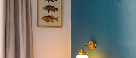 A bedroom with a bed, floral pillows, a wall-mounted lamp, and a framed fish illustration.