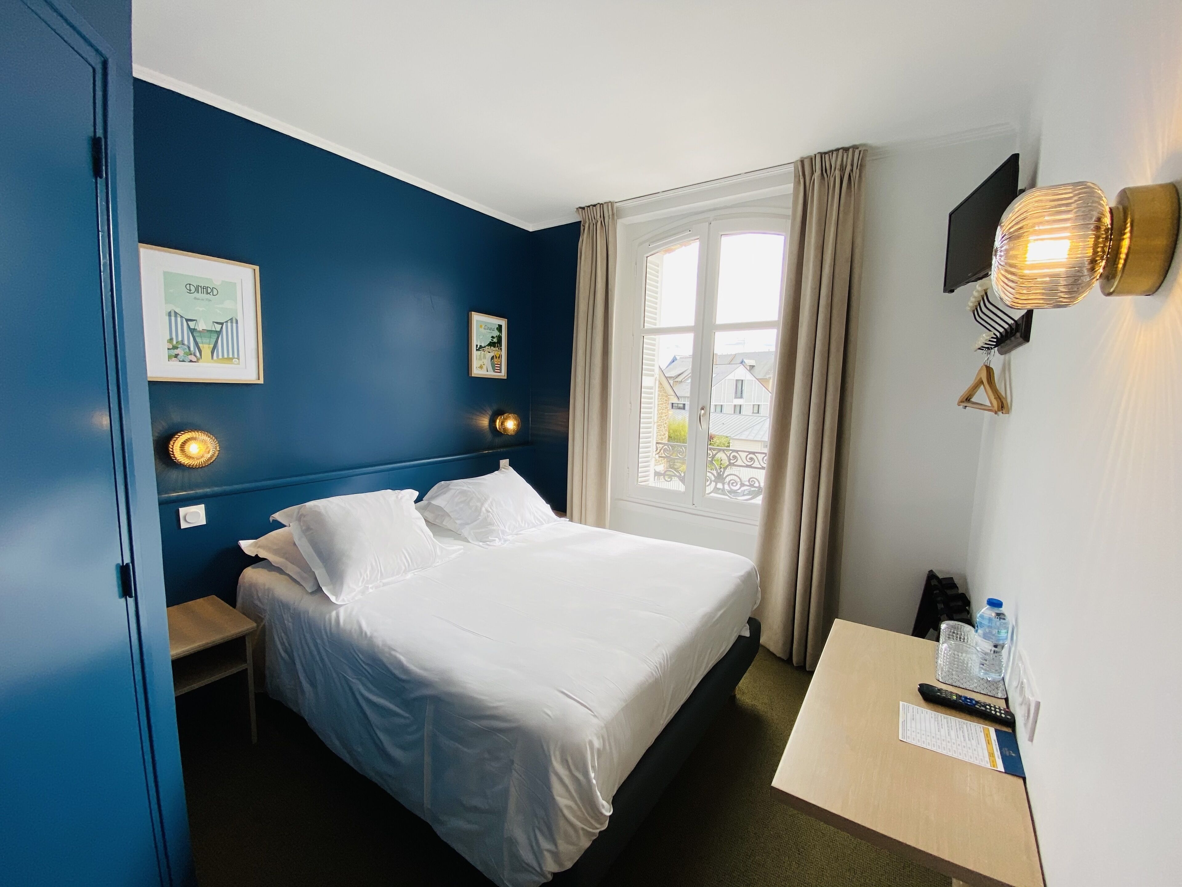Una habitación de hotel con una cama, mesitas de noche, un escritorio con silla, ventana con cortinas y luces en la pared.
