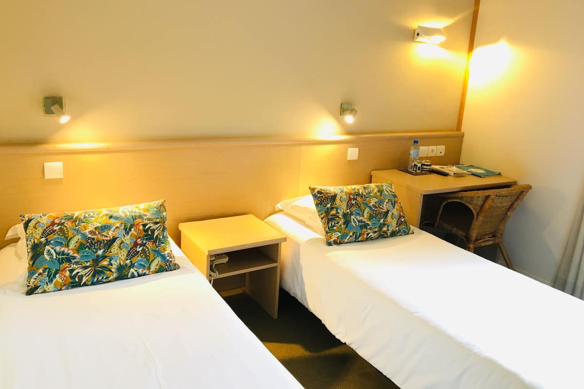 Habitación de hotel con dos camas individuales, cada una con un cojín floral de colores, una mesita de noche pequeña y una mesita auxiliar. Cuenta con un cabecero de madera, una lámpara de pared y un pequeño escritorio con una silla y una botella de agua.