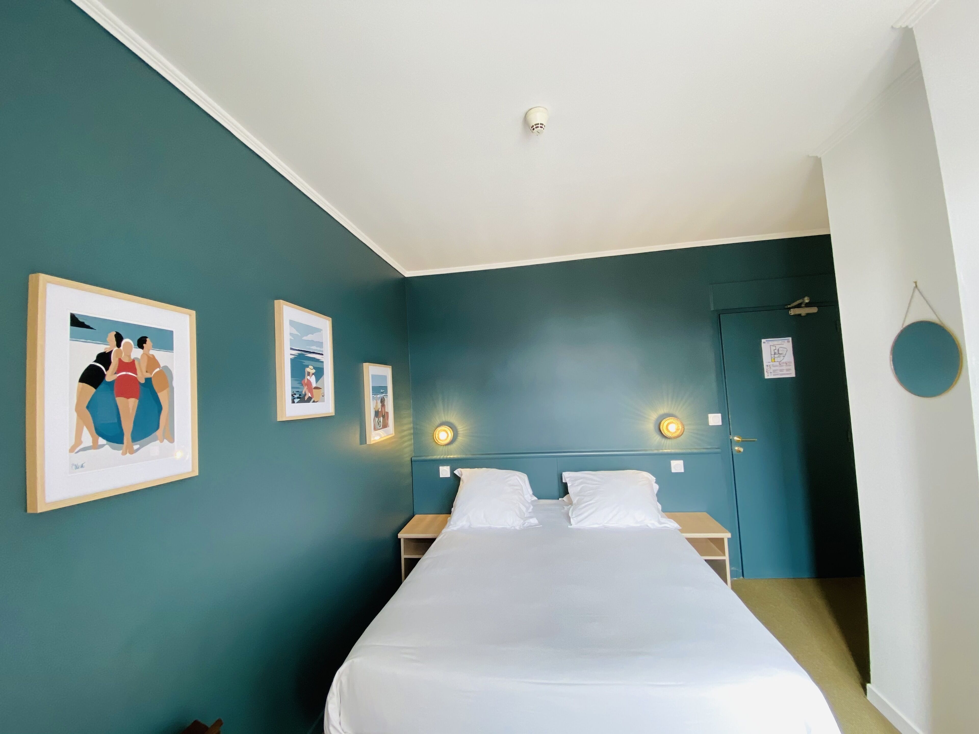 Una habitación de hotel con paredes color turquesa, una cama con ropa de cama blanca y tres cuadros en la pared.