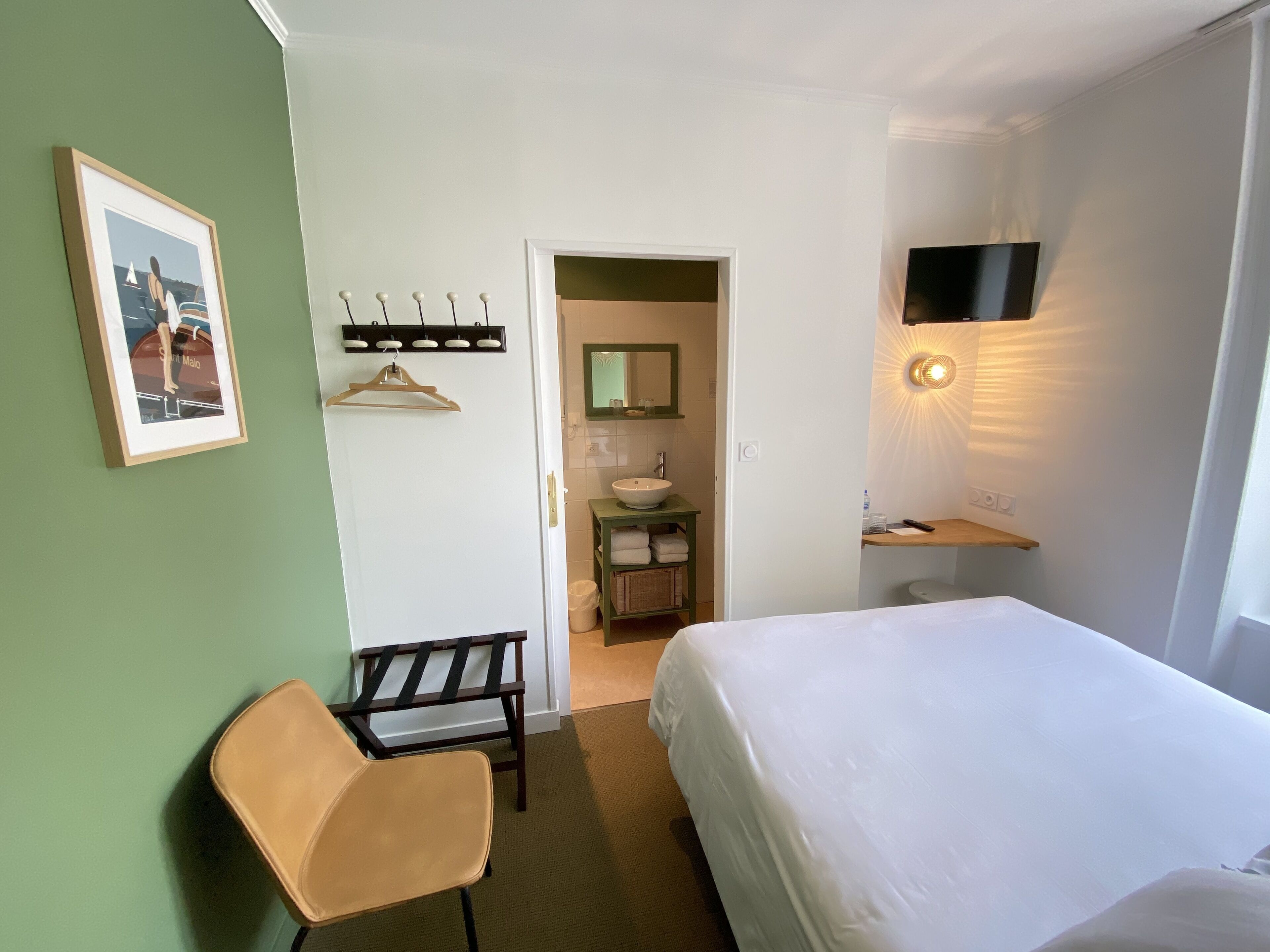 Habitación de hotel con cama, una silla, baño con lavamanos y espejo, y televisor montado en la pared.