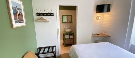 Habitación de hotel con cama, una silla, baño con lavamanos y espejo, y televisor montado en la pared.