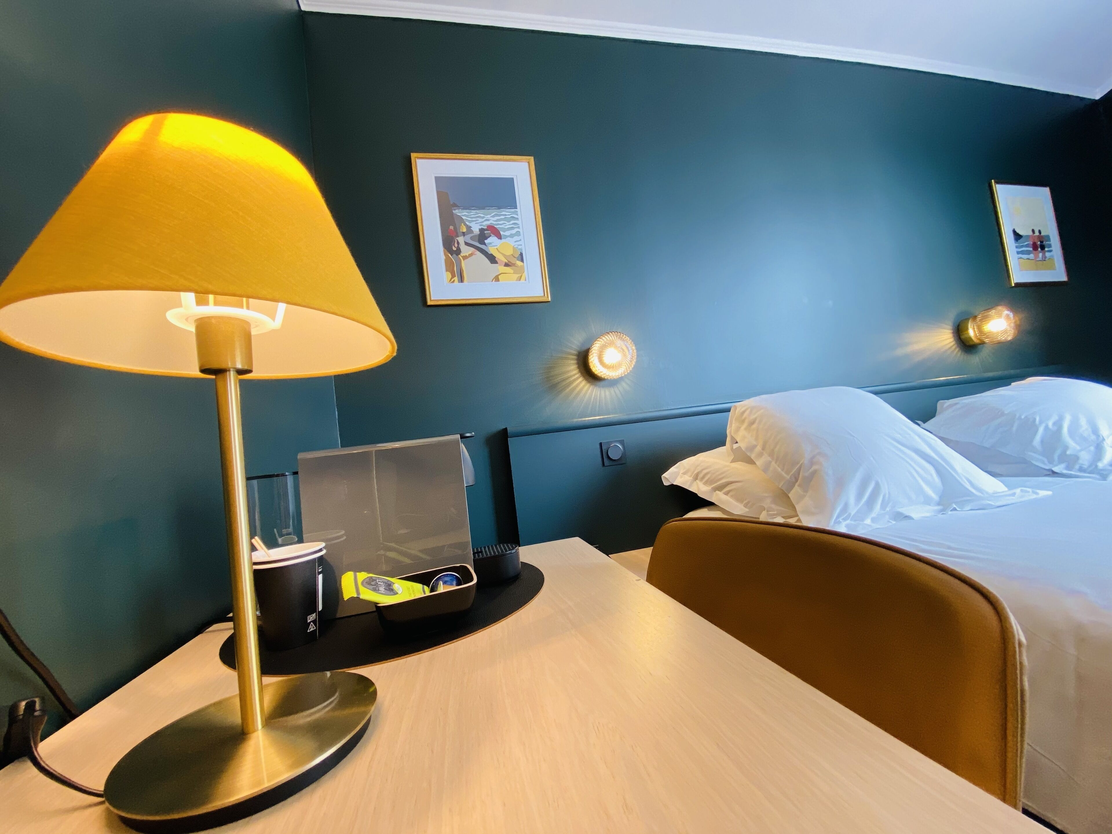 A hotel room with a bed, a desk with a lamp, a charging station, and artwork on the walls.