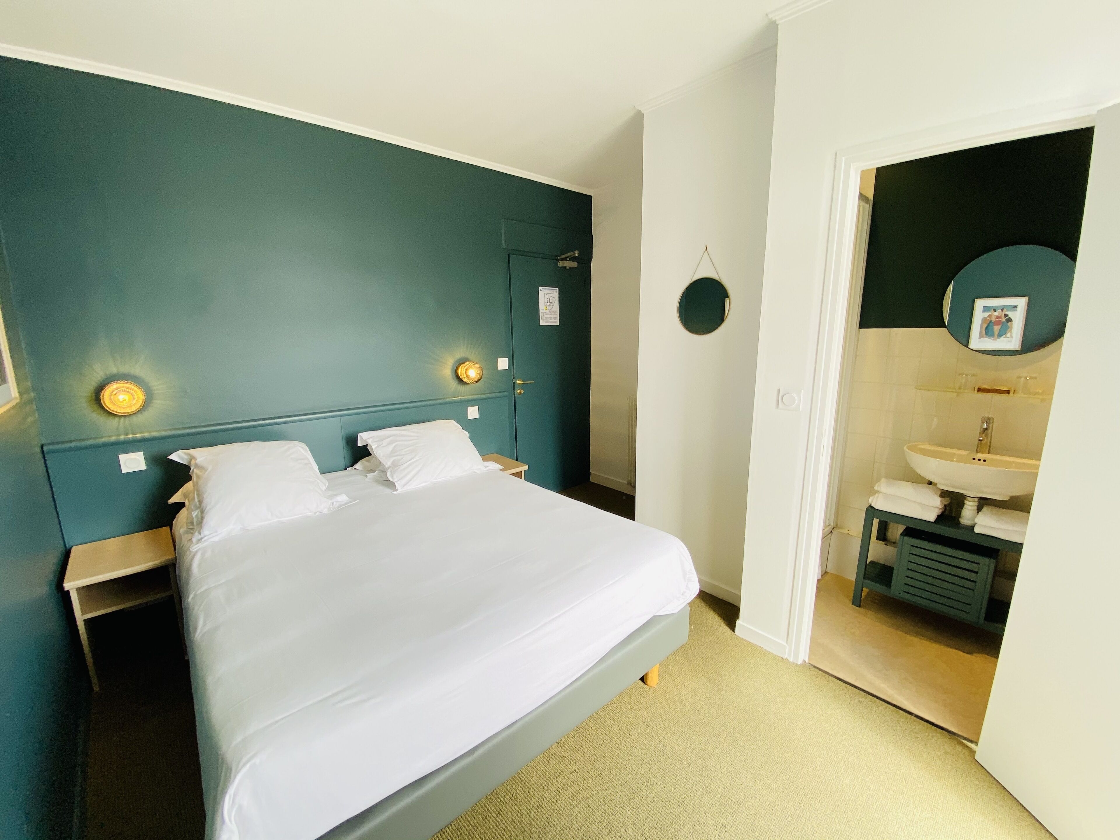 Une chambre à coucher comprenant un lit, des tables de chevet, une lampe fixée au mur et une porte donnant sur une salle de bain équipée d’un lavabo et d’un miroir.