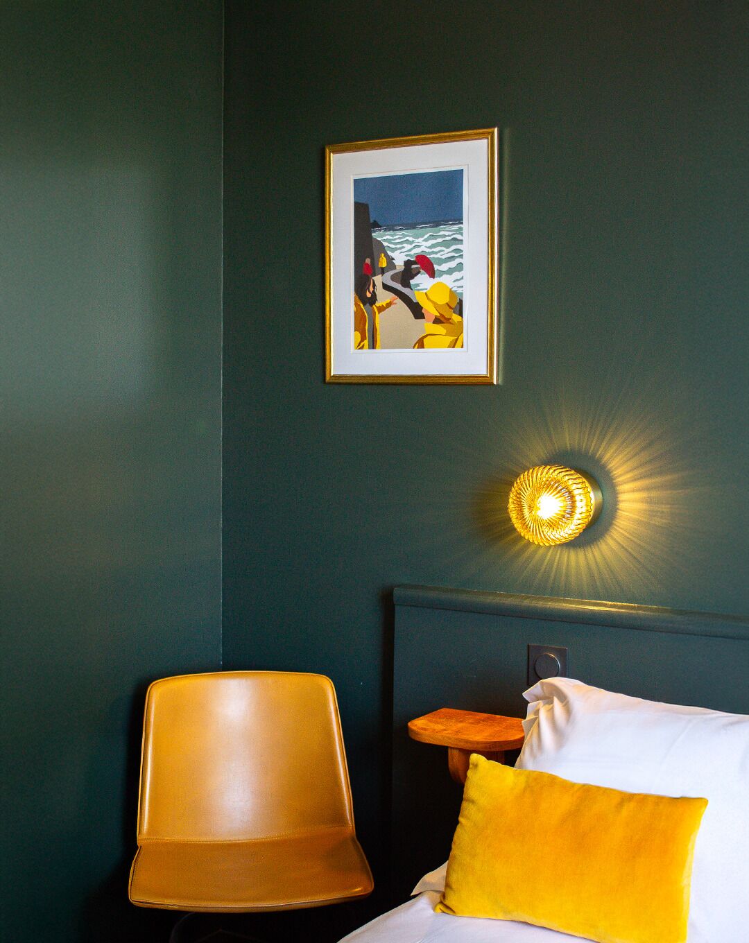 Quarto de hotel com parede verde escura, quadro com cena de praia, cadeira moderna, mesa de cabeceira com abajur e cama com travesseiro amarelo.