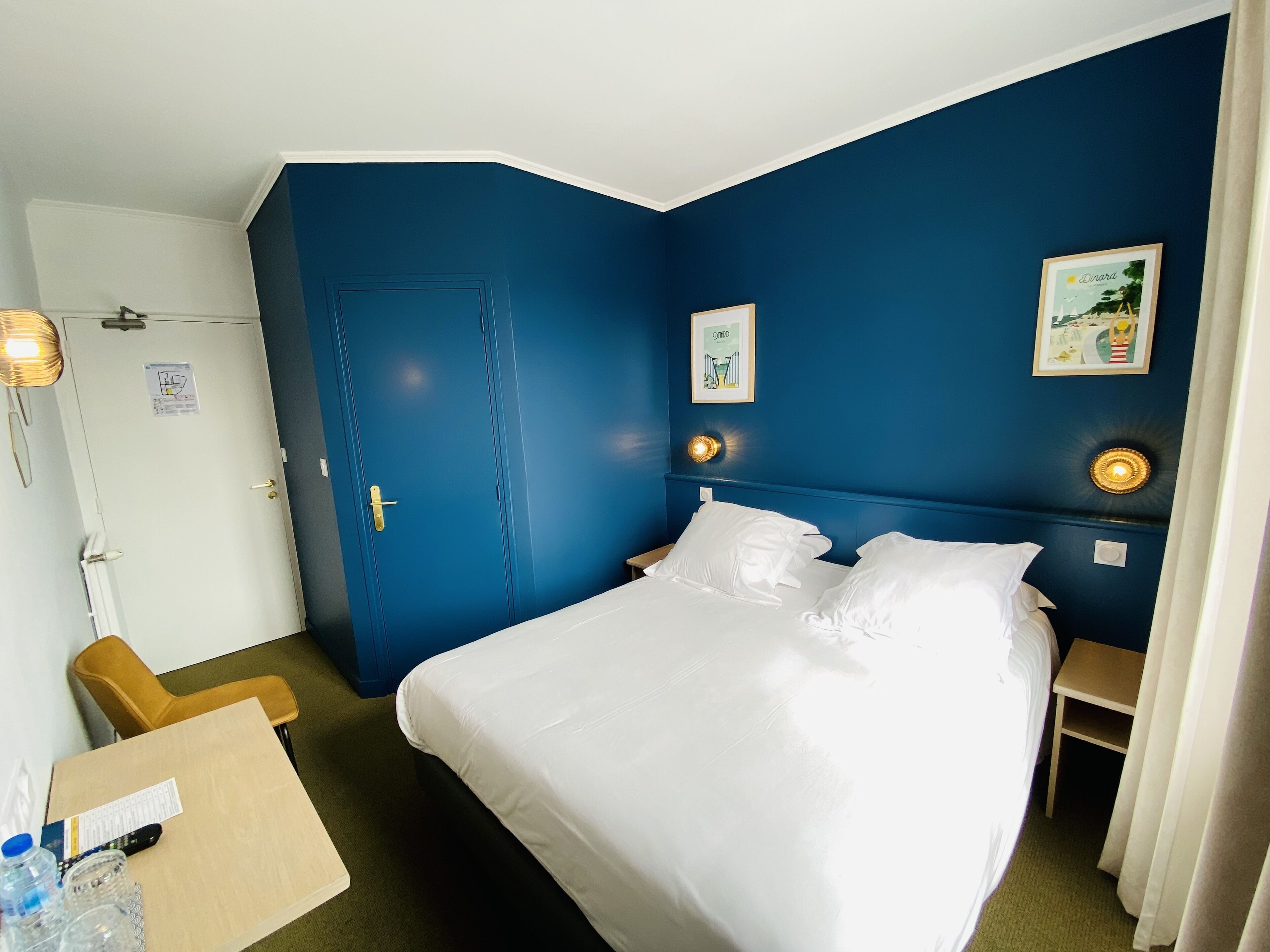 A hotel room with a large bed, a desk with a chair, a door, and two wall-mounted light fixtures.