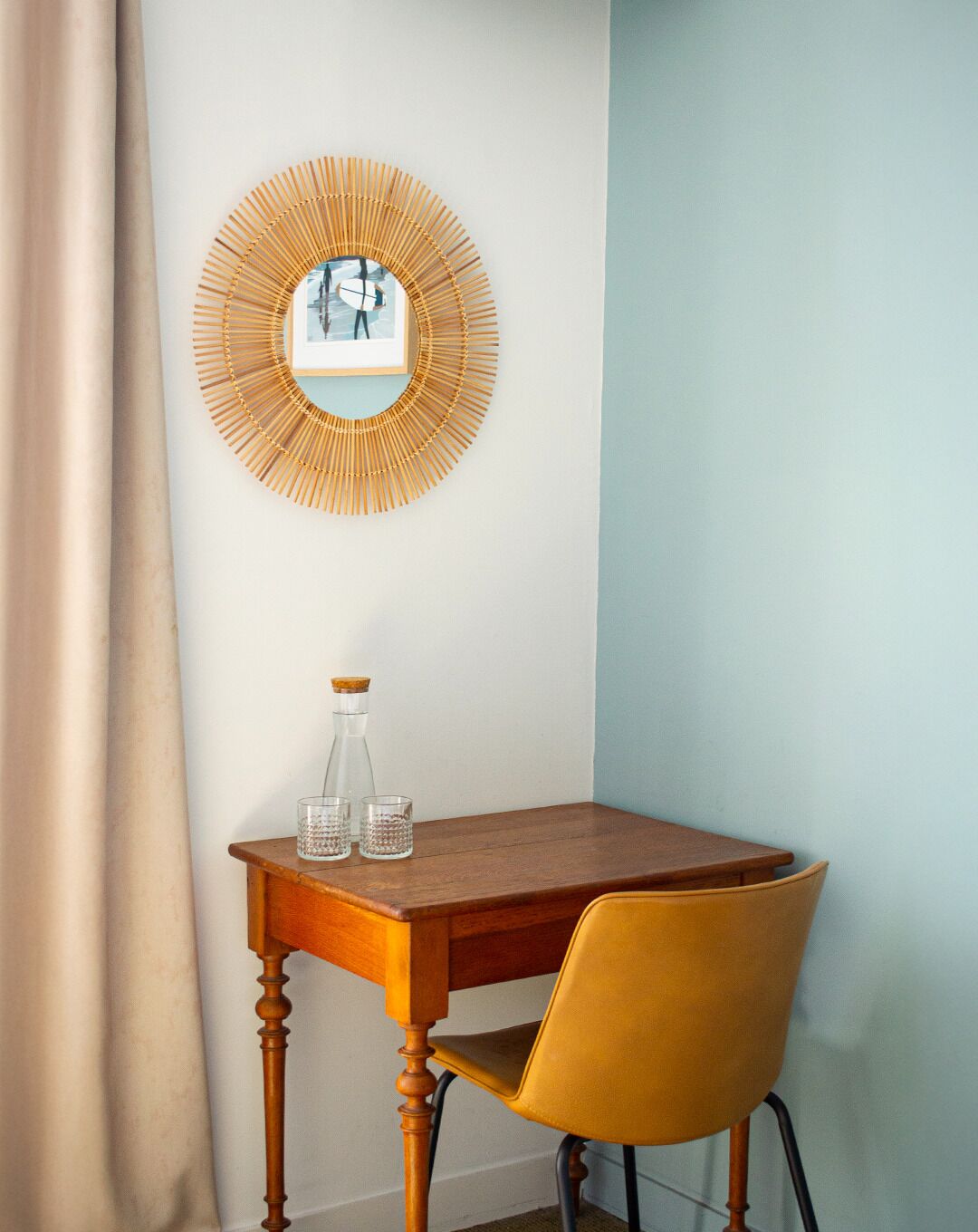 Ein Zimmer mit Holztisch, gelbem Stuhl, wandmontiertem Spiegel und einem Fenster mit Vorhang.