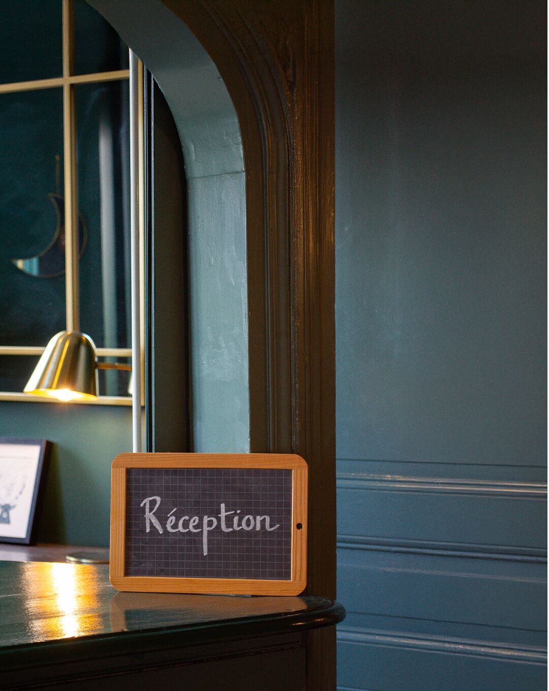 Une enseigne indiquant le service d’accueil, avec le mot « Accueil » inscrit sur un fond quadrillé, placée sur une table près d’une fenêtre, à côté d’une lampe.