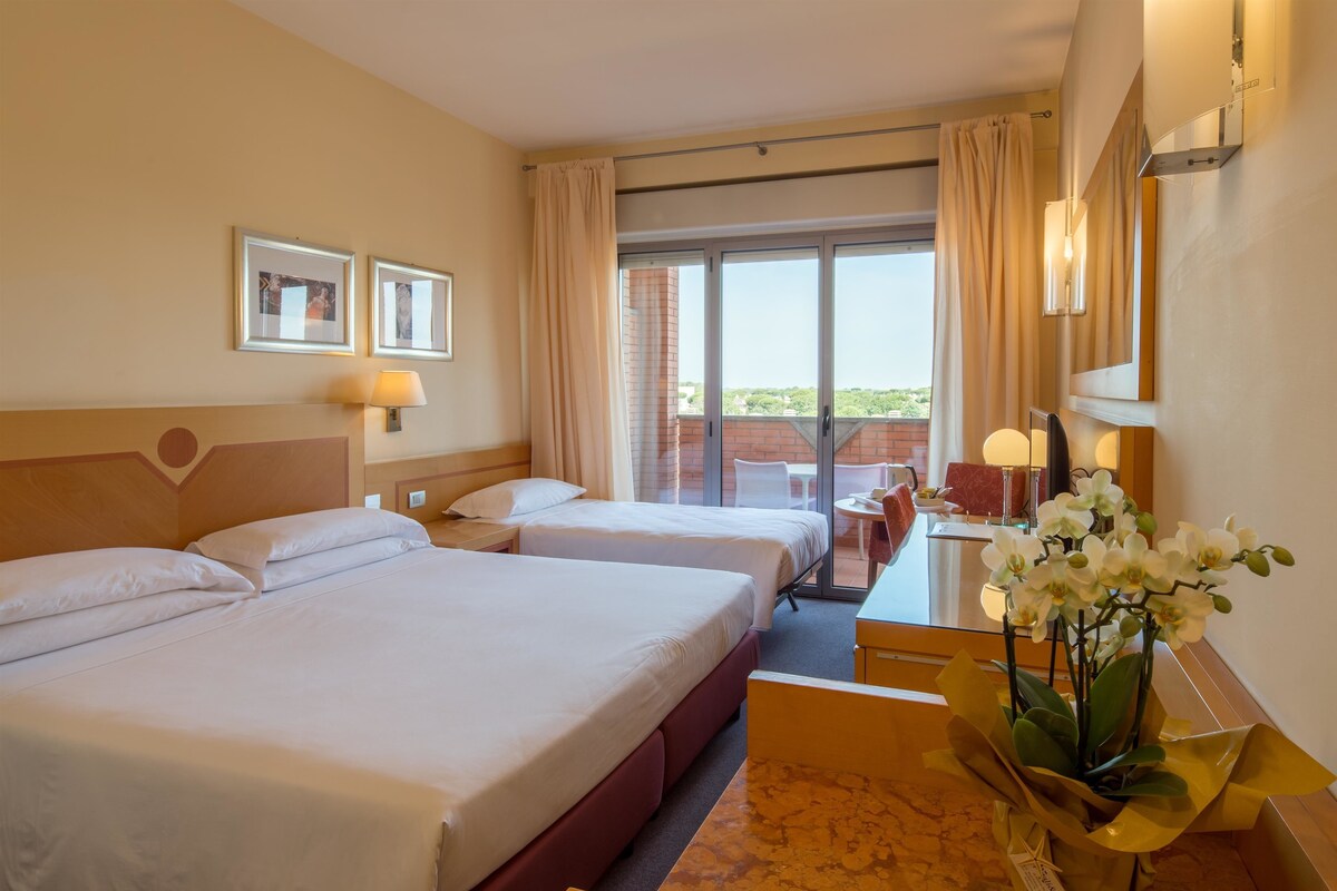 A hotel room with two beds, a desk, a chair, a small table, a vase of flowers, and a balcony with a view.
