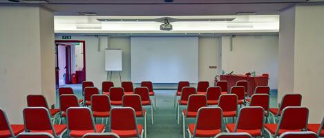 A conference room with rows of red chairs, a whiteboard, and a projector on the ceiling.