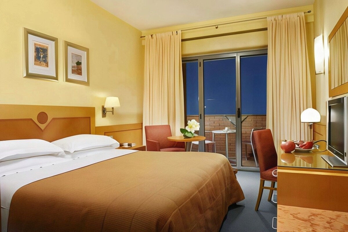 A hotel room with a large bed, two chairs, a small table with a flower, and a TV.