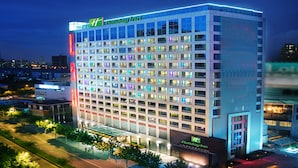 Un hôtel Holiday Inn à plusieurs étages, doté d’une façade moderne en verre, de fenêtres illuminées et d’une grande enseigne « Holiday Inn » sur le toit.