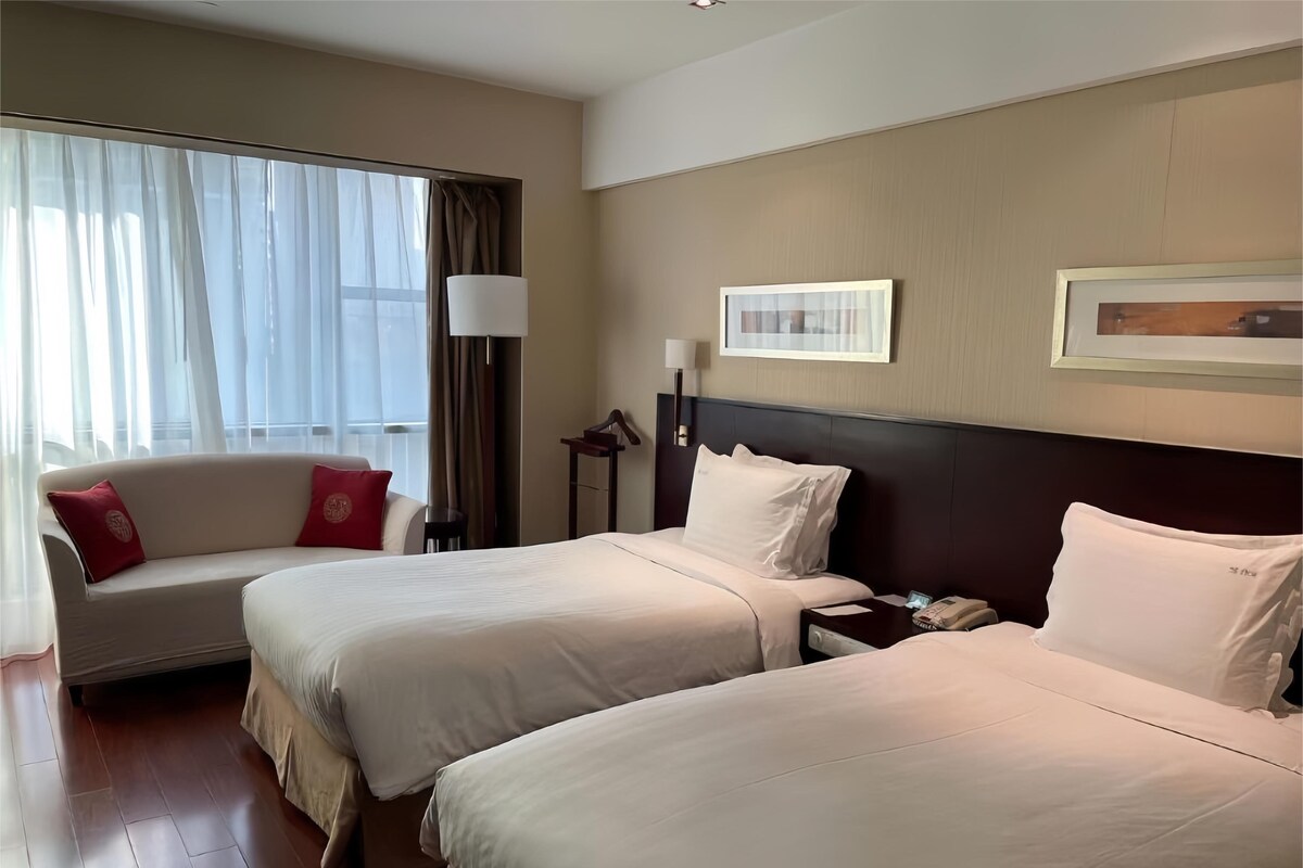 A hotel room with two beds, a sofa, a bedside table with a lamp, and a framed picture on the wall.