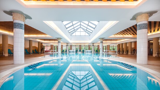 A large indoor swimming pool with a geometric tile design, surrounded by columns and a skylight ceiling. - Hotel Aquarell (Cegled)