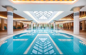 A large indoor swimming pool with a geometric tile design, surrounded by columns and a skylight ceiling. - Hotel Aquarell (Cegled)