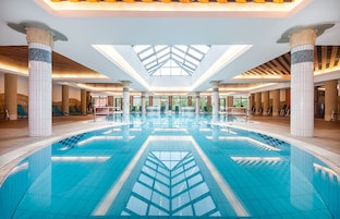 A large indoor swimming pool with a geometric tile design, surrounded by columns and a skylight ceiling.