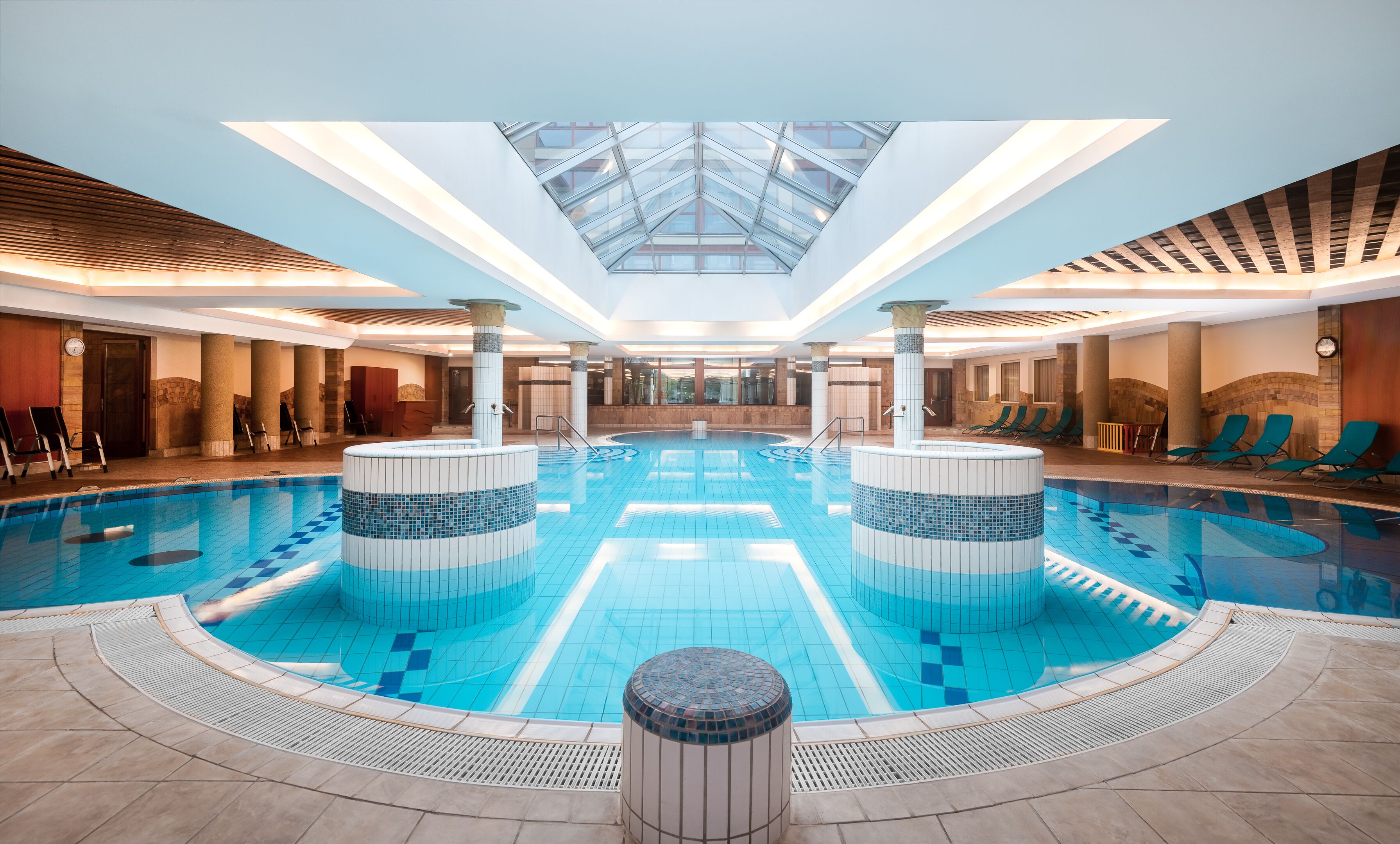 A large indoor swimming pool with a skylight ceiling, lounge chairs, and a central column.