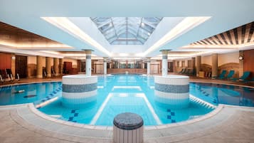 A large indoor swimming pool with a skylight ceiling, lounge chairs, and a central column.