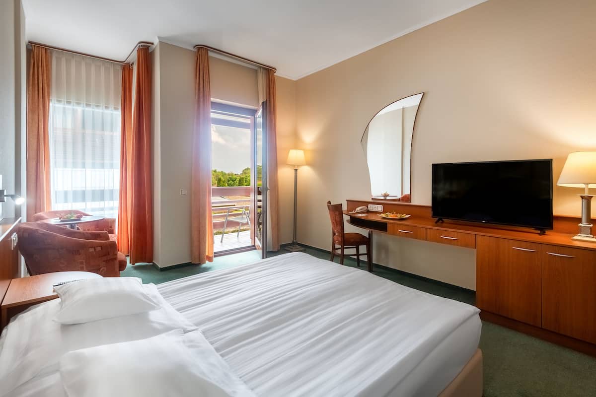 A hotel room with a bed, a desk with a television, a chair, and a desk lamp.