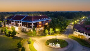 A large building with a red roof and multiple windows, surrounded by greenery and a well-lit street. - Hotel Aquarell (Cegled)
