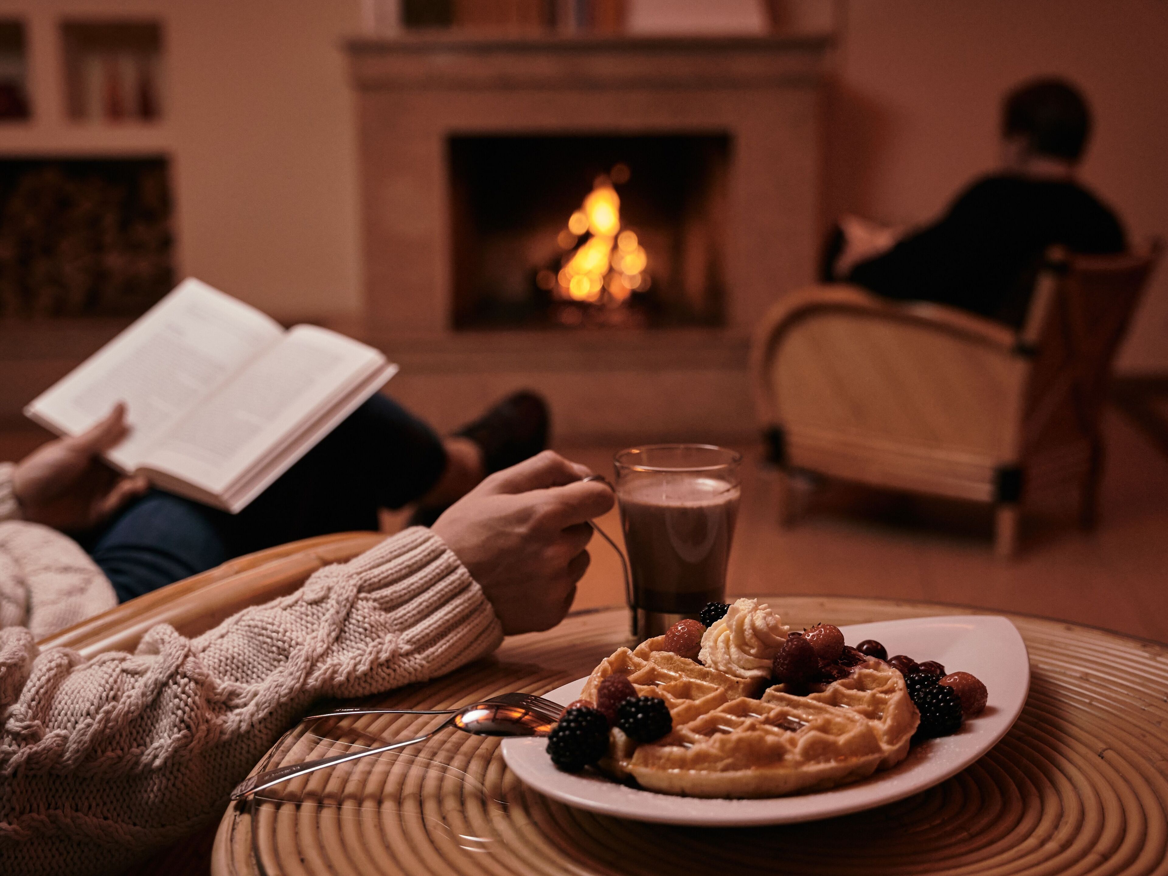Una habitación acogedora con chimenea, un plato de gofres con frutos rojos y crema batida, y una persona leyendo un libro.