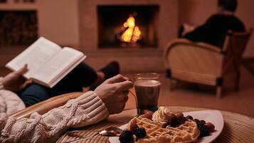 Una habitación acogedora con chimenea, un plato de gofres con frutos rojos y crema batida, y una persona leyendo un libro.