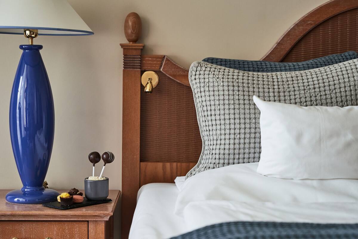 Una cama bien hecha con almohadillas estampadas, una mesita de noche de madera con una lámpara azul y una pequeña bandeja con bombones.