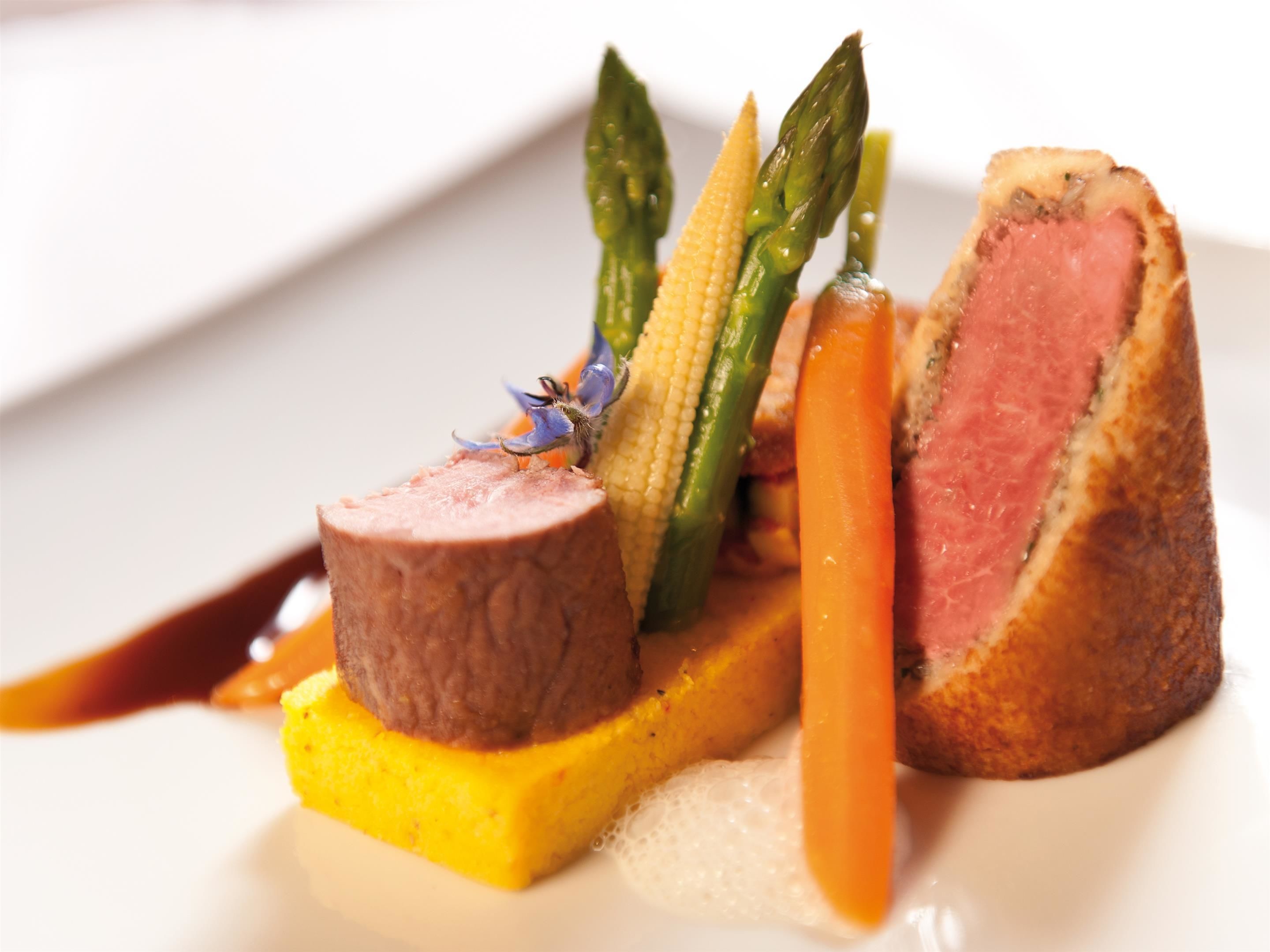 A plated dish with a piece of meat, asparagus, corn, and a yellow puree.