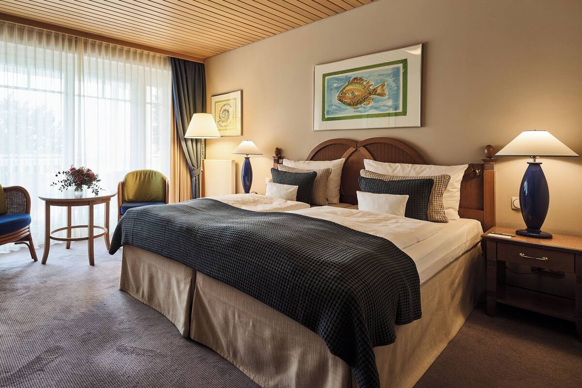 Habitación de hotel con una cama grande, dos mesitas de noche con lámparas, una pequeña mesa con un florero y una silla.