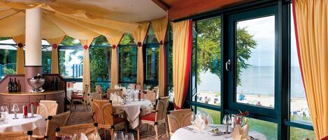 A restaurant with tables set for dining, large windows with curtains, and a view of the beach.