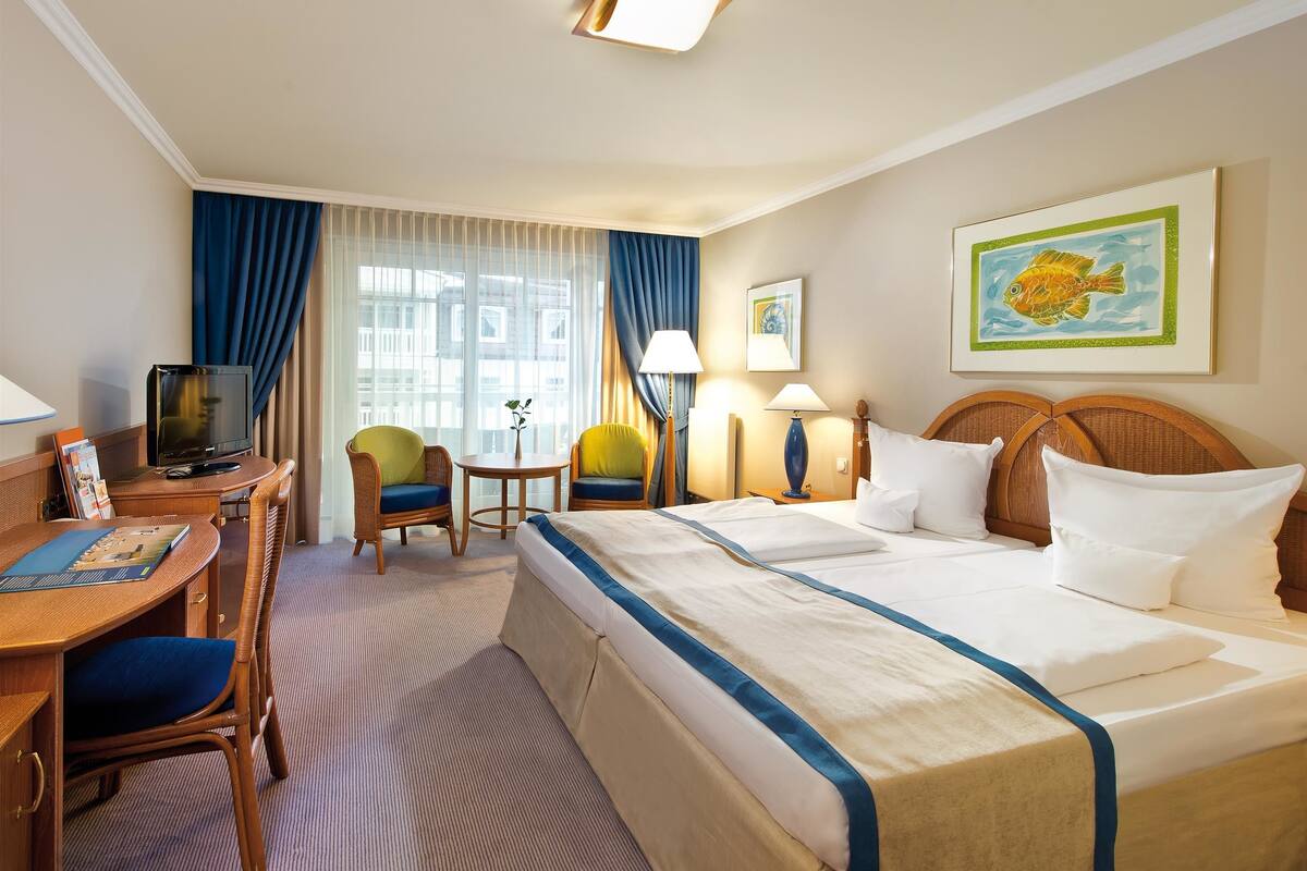 Una habitación de hotel con cama, escritorio con silla, televisor y ventana con cortinas.