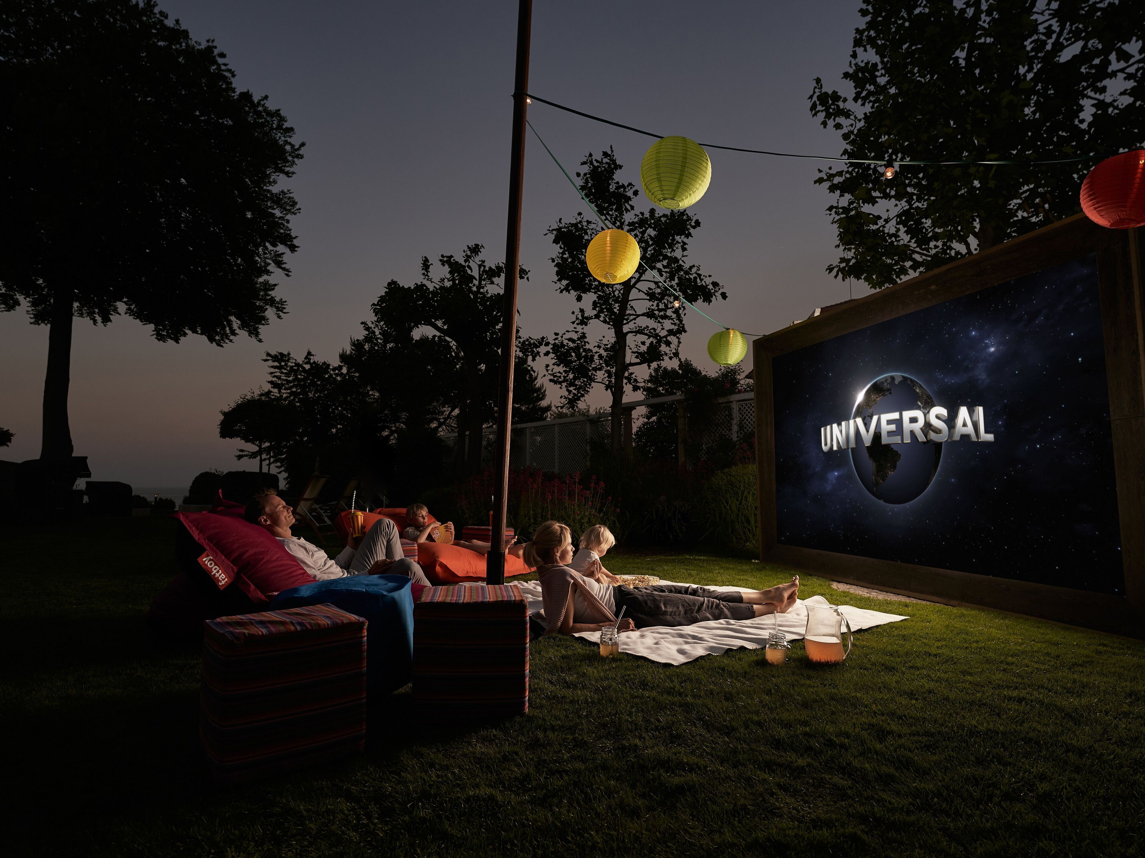 Ulkoilmaelokuvanäytös, taustalla Universal-yhtiön logo, ihmiset loikoilevat ruohikolla ja yläpuolella on valonauhoja.