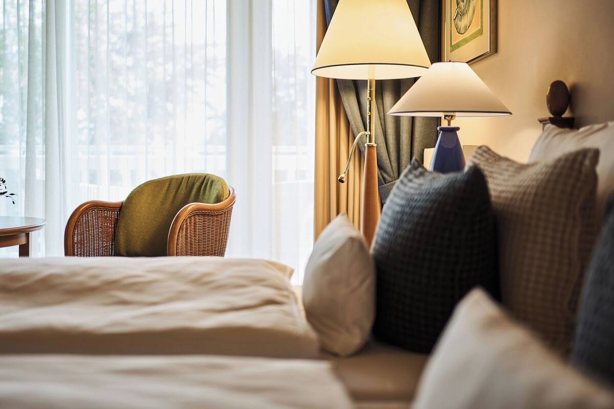 Una habitación de hotel con cama, una silla, dos lámparas y un ventanal con cortinas.