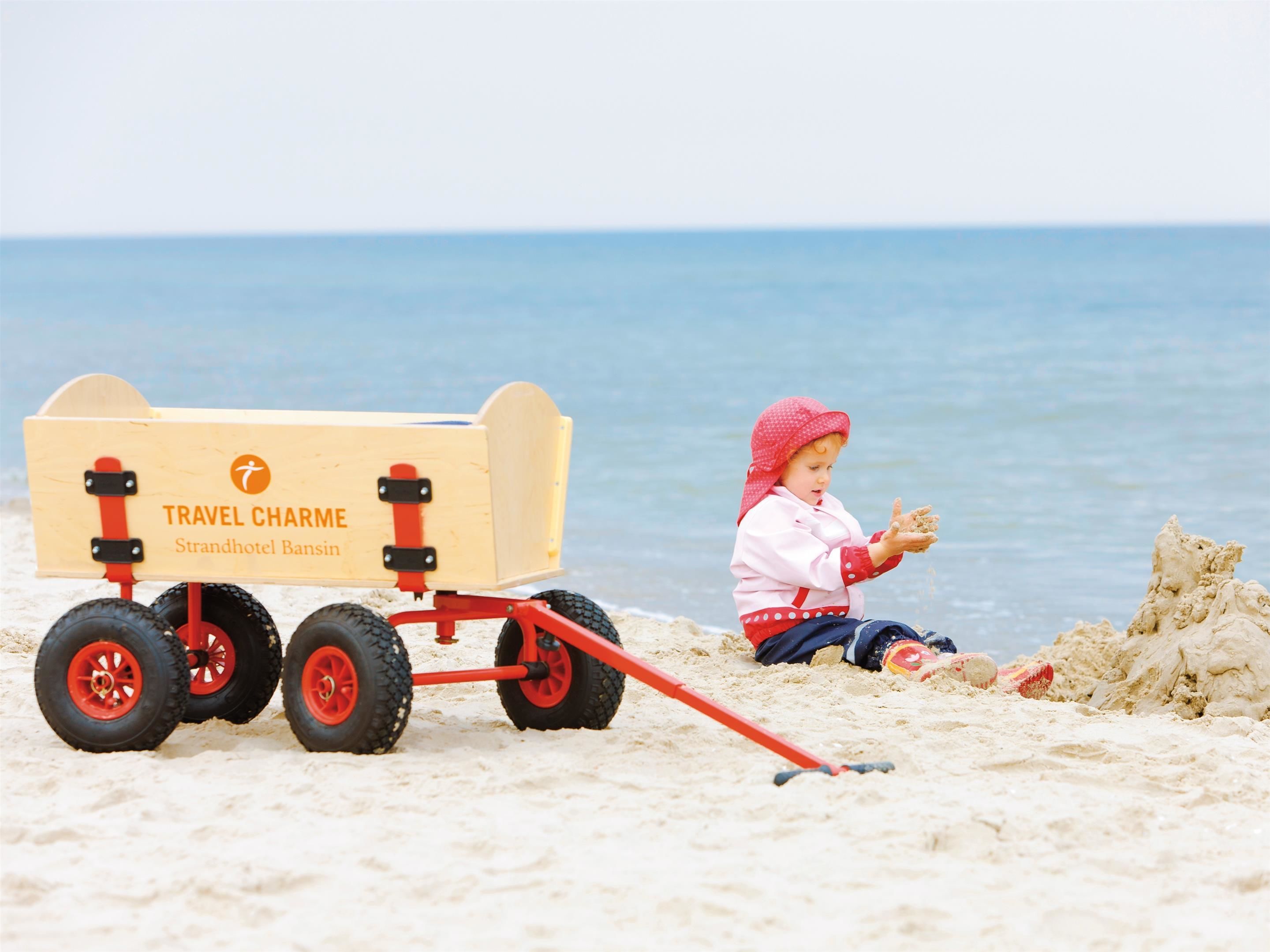 Niño jugando con arena en la playa, junto a un carro de madera con la leyenda 'TRAVEL CHARME Strandhotel Bansin'.