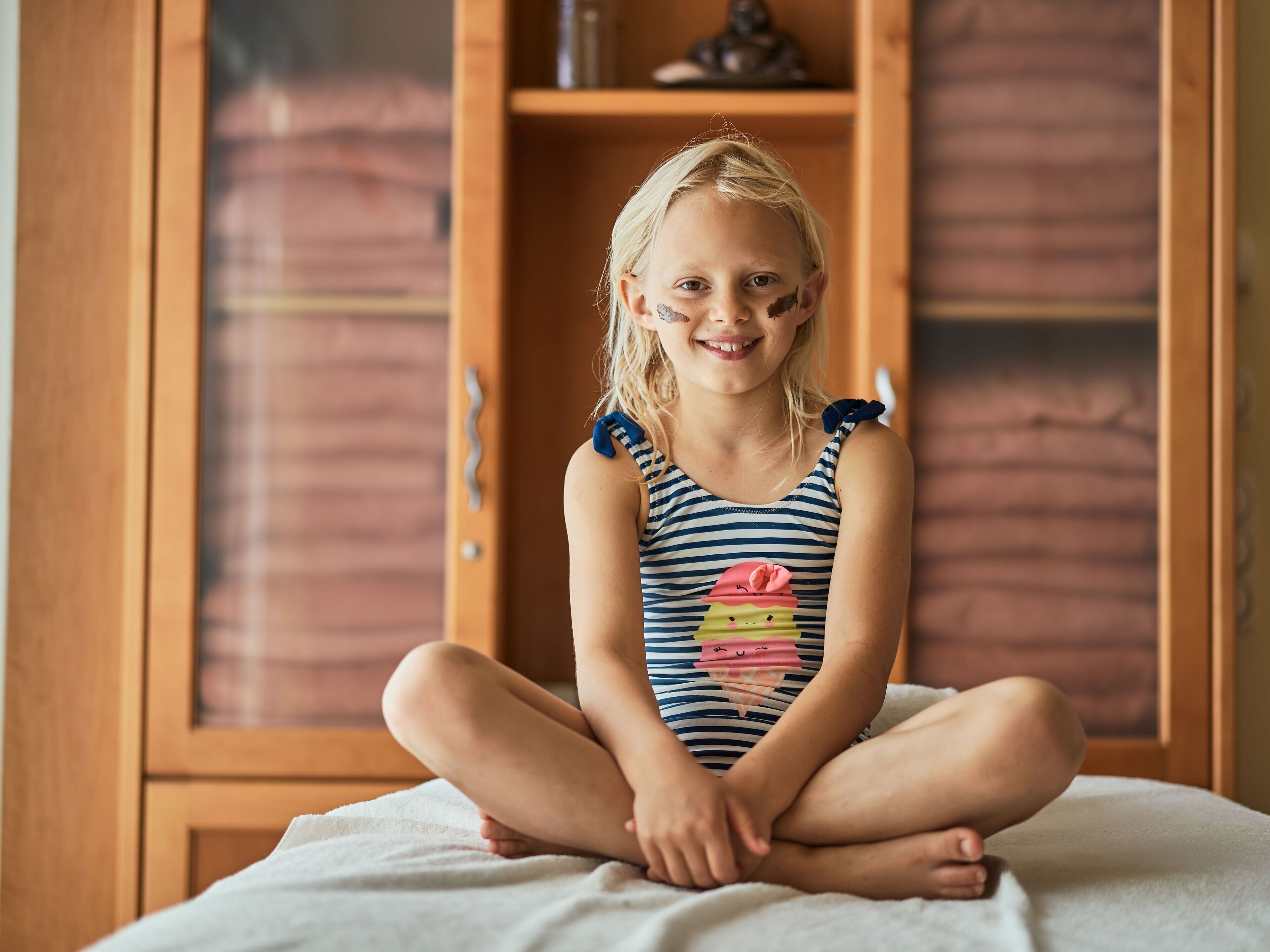 A child sitting on a bed in a room with wooden furniture and a cabinet.