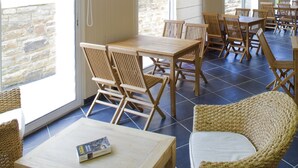 A restaurant with wooden tables and chairs, a wicker chair, and a tiled floor. - Résidence Le Domaine des Glénan (Fouesnant)