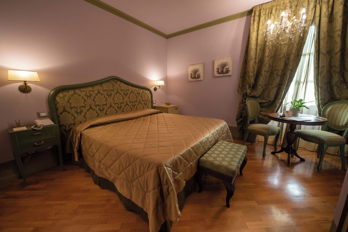 Un dormitorio con una cama grande, mesitas de noche, una pequeña mesa con sillas y una ventana con cortinas.