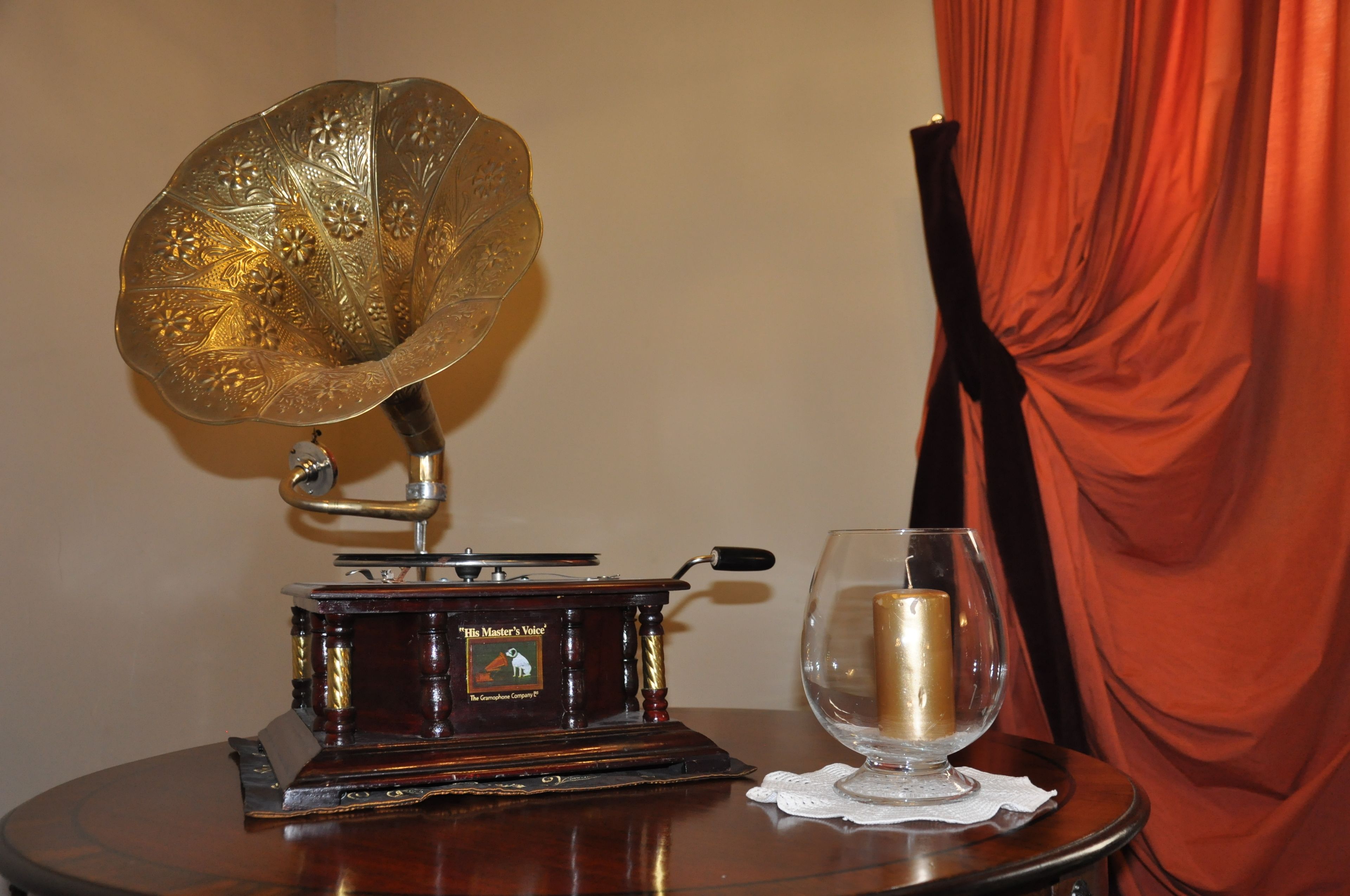 Ein Grammophon aus vergangenen Zeiten auf einem Holztisch neben einem Glas mit Kerze, vor einem roten Vorhang und einer schlichten Wand.