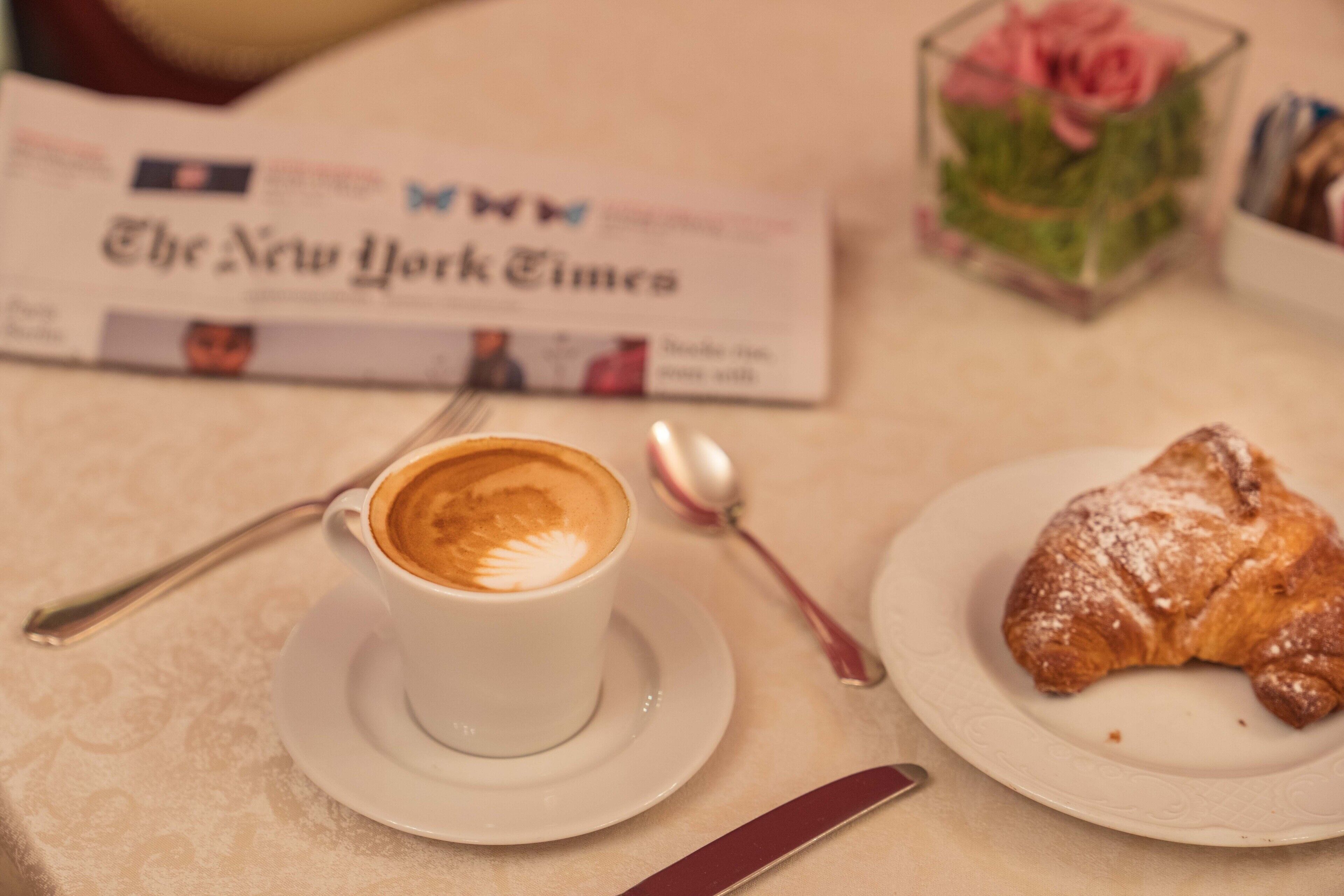 Una mesa con una taza de café, un croissant, una cuchara, un cuchillo y un periódico.
