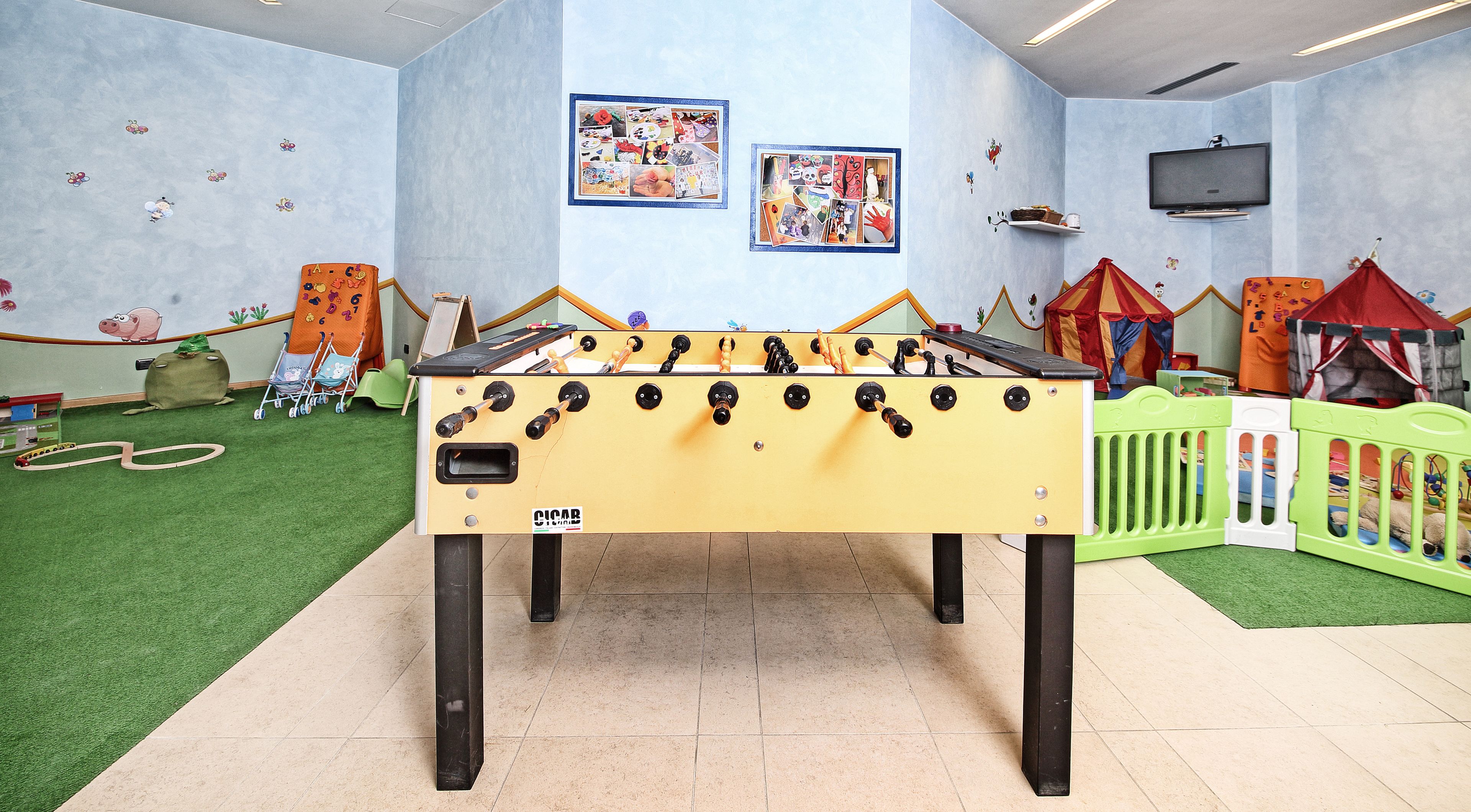 Une table de baby-foot dans une salle de jeux, avec un tapis d’herbe artificielle verte, une maisonnette et des dessins d’enfants accrochés aux murs.