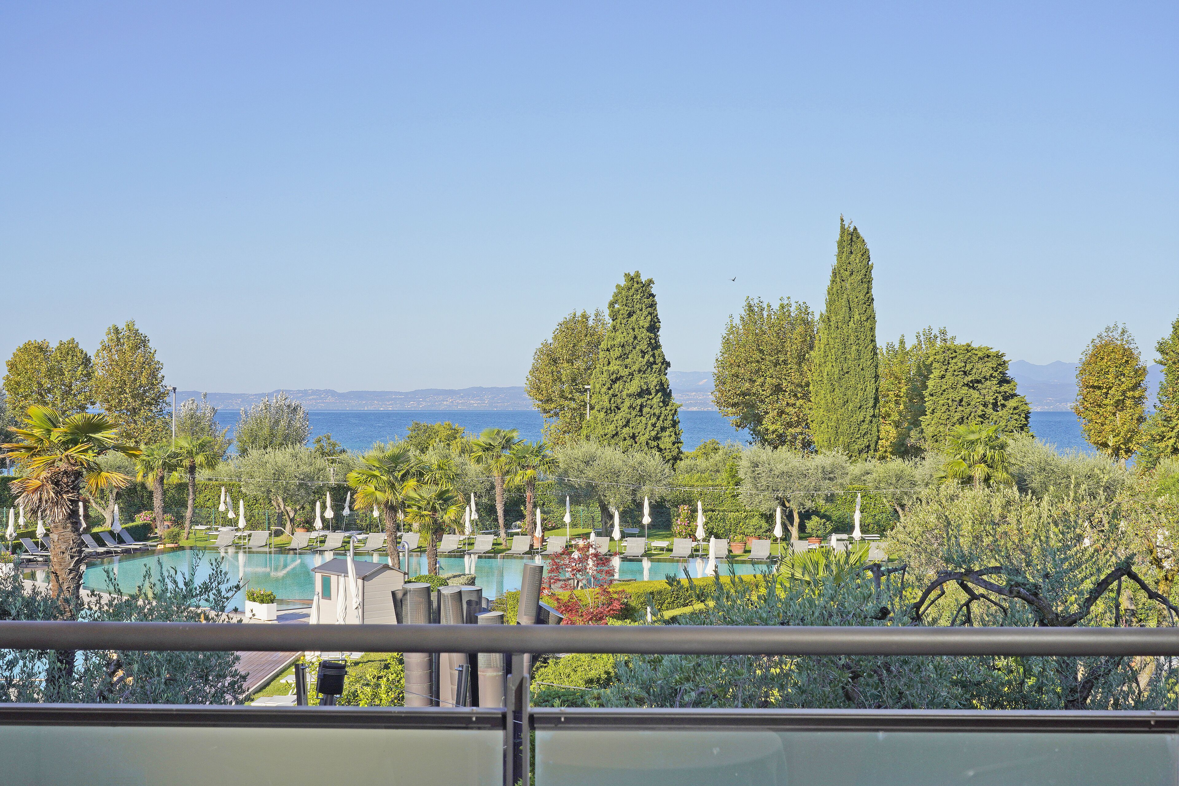 Zona de piscina con sillones y sombrillas, rodeada de árboles y vista al mar.