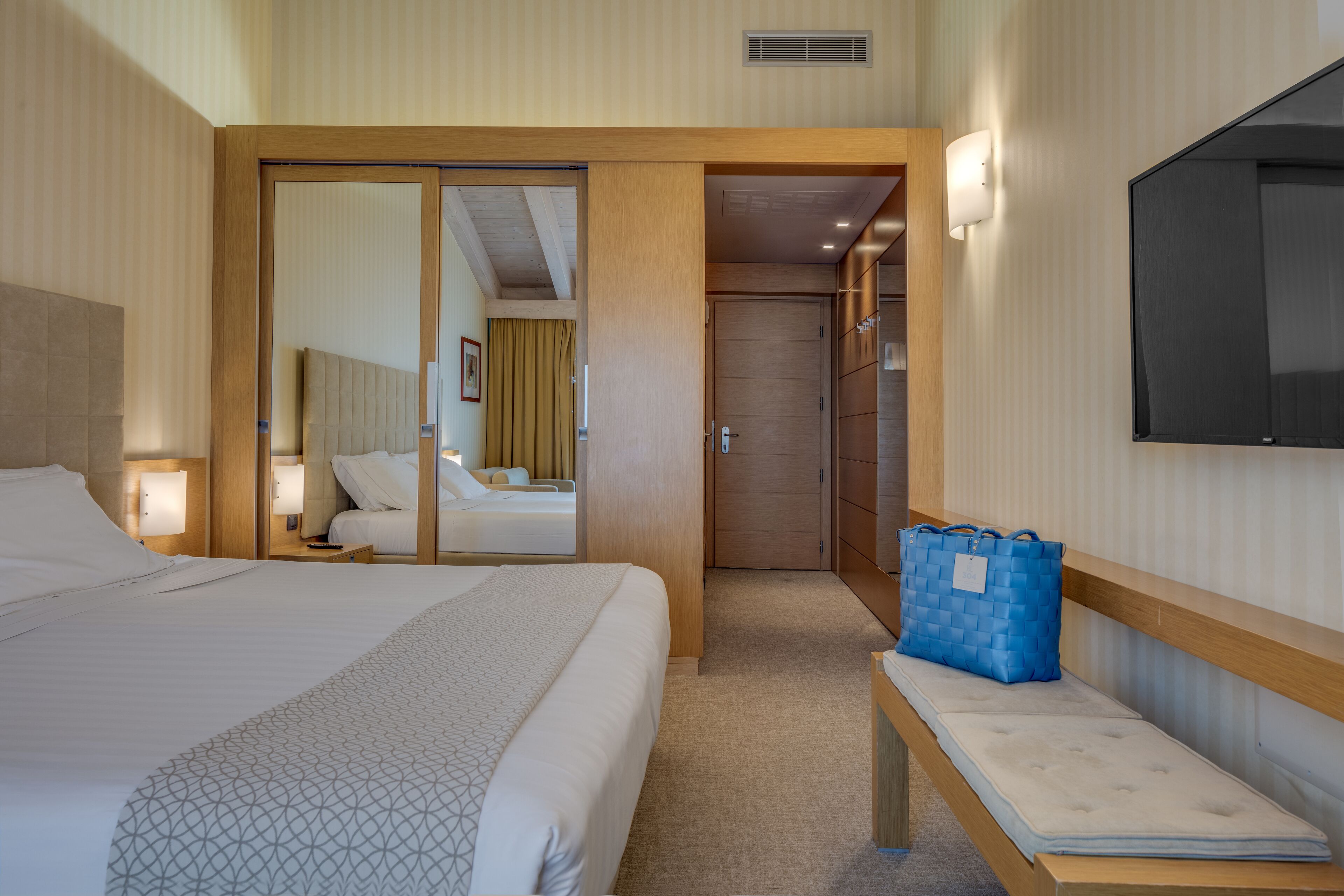 A hotel room with a large bed, a bedside table with a lamp, a blue bag on a bench, and a TV mounted on the wall.