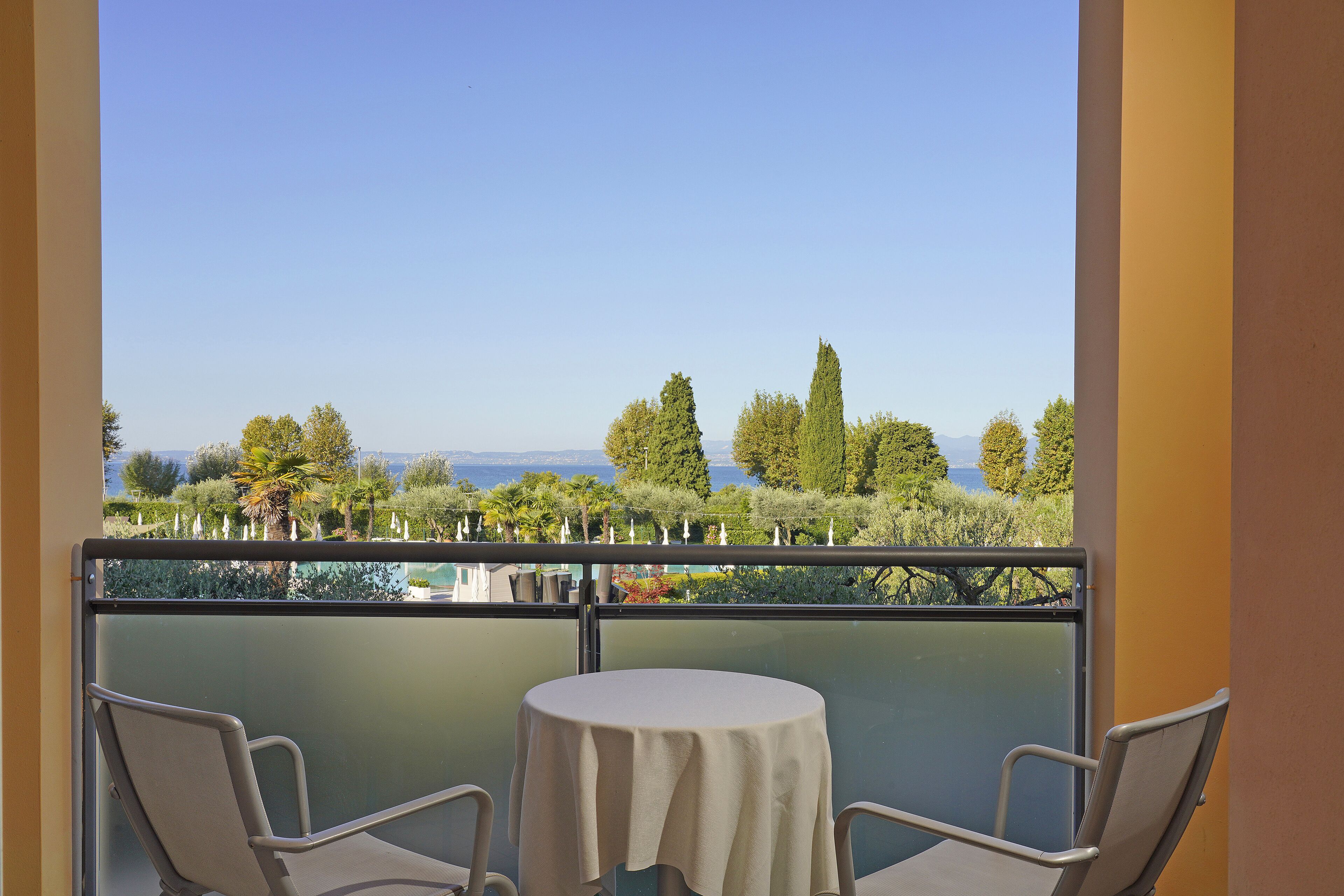 Un balcon avec une table et deux chaises, donnant sur un paysage pittoresque avec des arbres et une étendue d’eau.