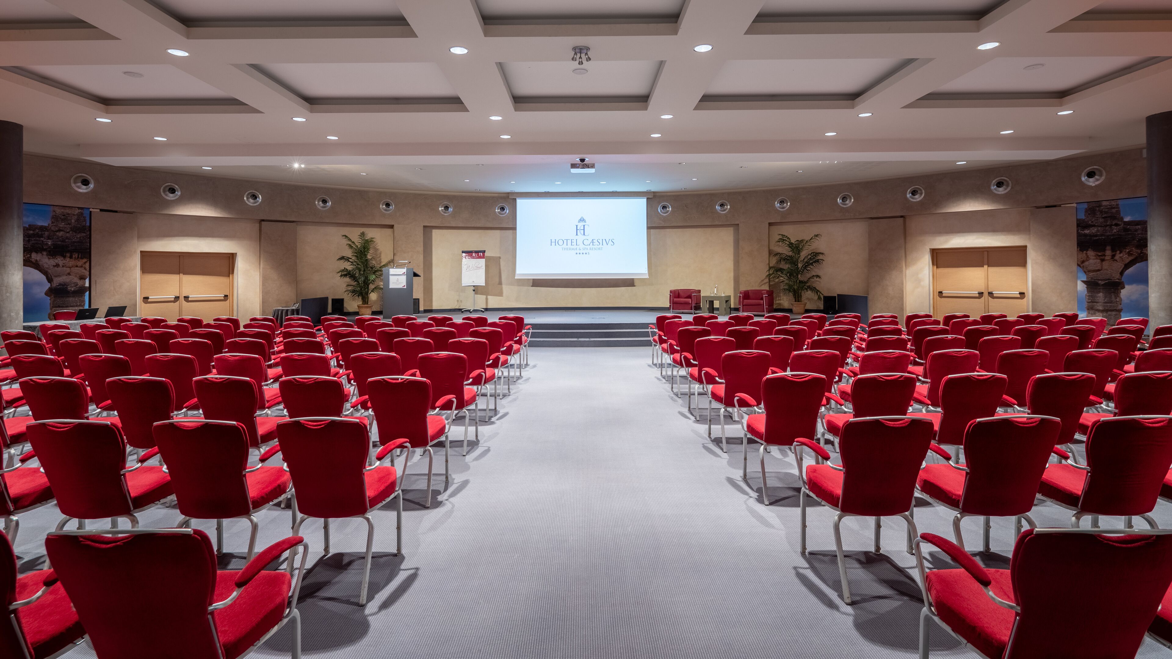 A conference room with rows of red chairs, a stage with a screen displaying 'HOTEL CASIFS', and a modern ceiling design.