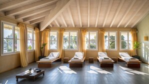 A sunlit room with wooden beams, large windows with curtains, and multiple sunbeds.
