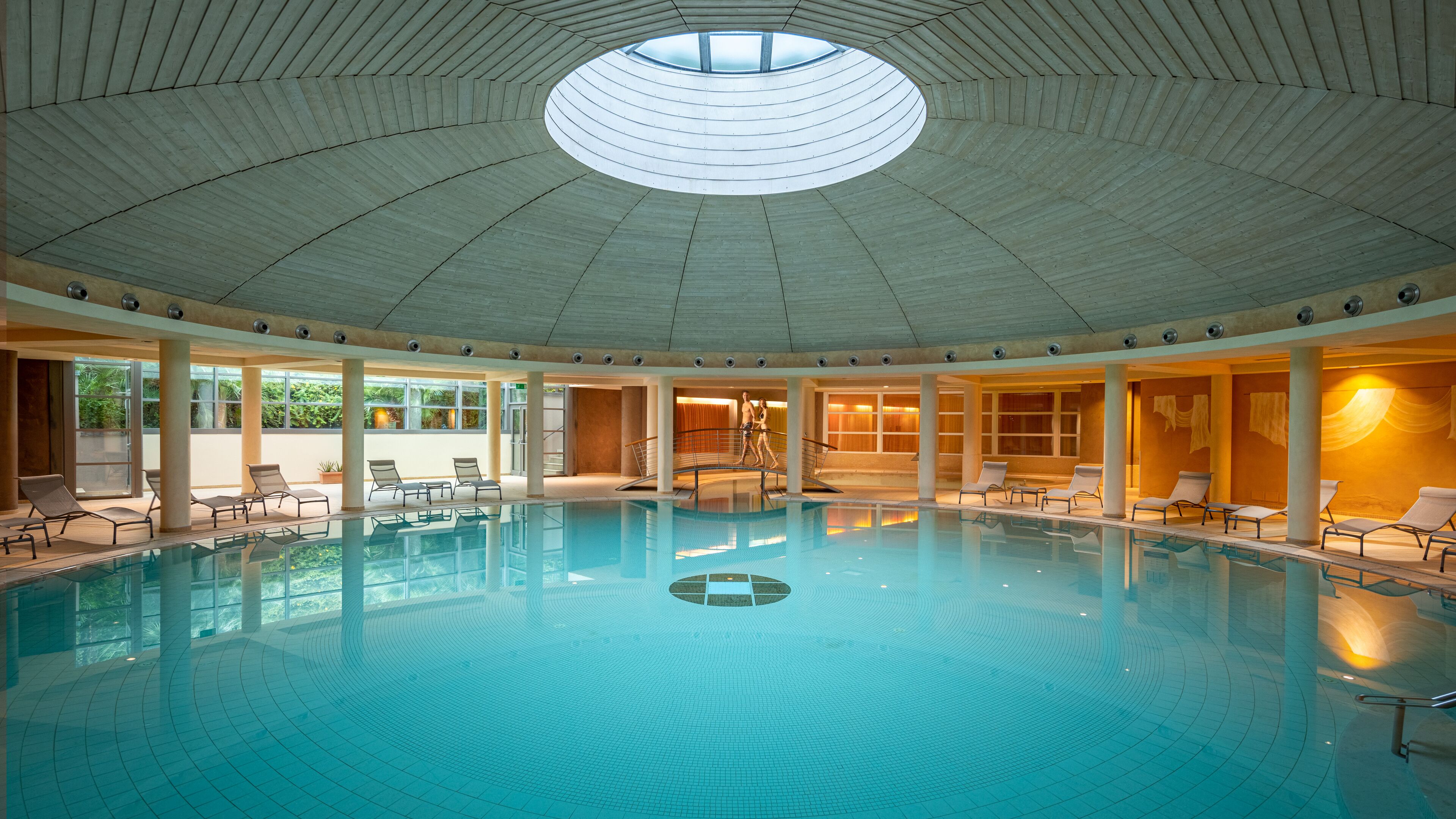 En stor indendørs swimmingpool med cirkulært tagdesign, omgivet af liggestole og et solbelyst område med store vinduer.