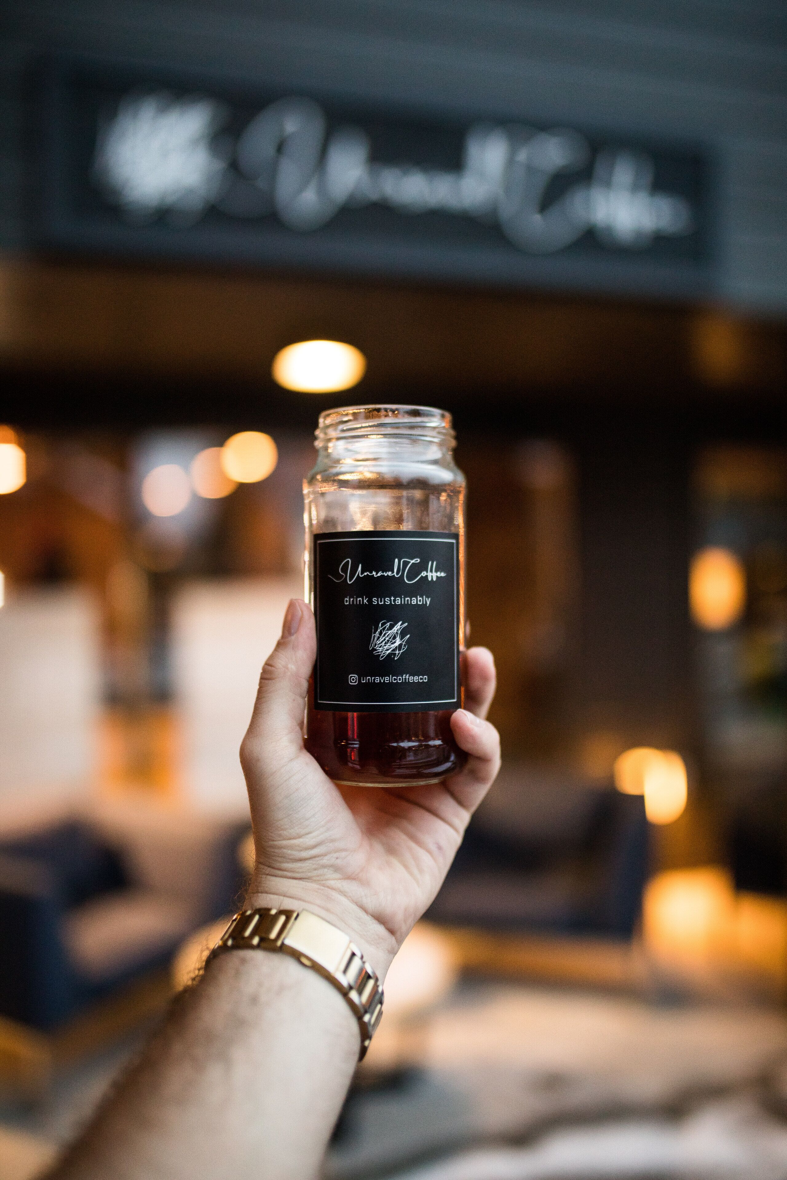 A hand holding a jar labeled "Unravelt Coffee" with a blurred background of a cafe interior.